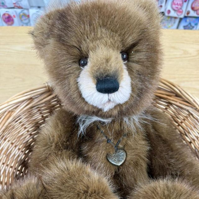 Front view of Charlie Bears Boggle sitting in wicker basket with heart pendant necklace