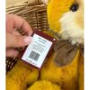 Hand holding tag of Charlie Bears Mrs Jones bear with brown bow and yellow plush fur in basket